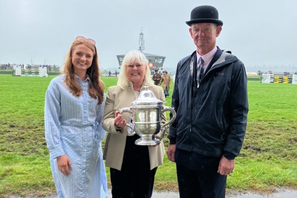 Two women and a man standing together with a silver trophy by a horse racing track