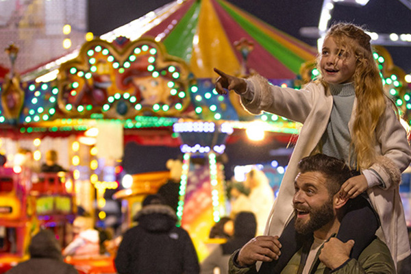 A Pointing Child Sitting On an Adults Shoulders with a Fun Fair Backdrop