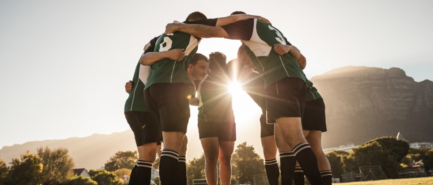 Group of male rugby players in a huddle on the pitch with the sun's rays shining through their arms